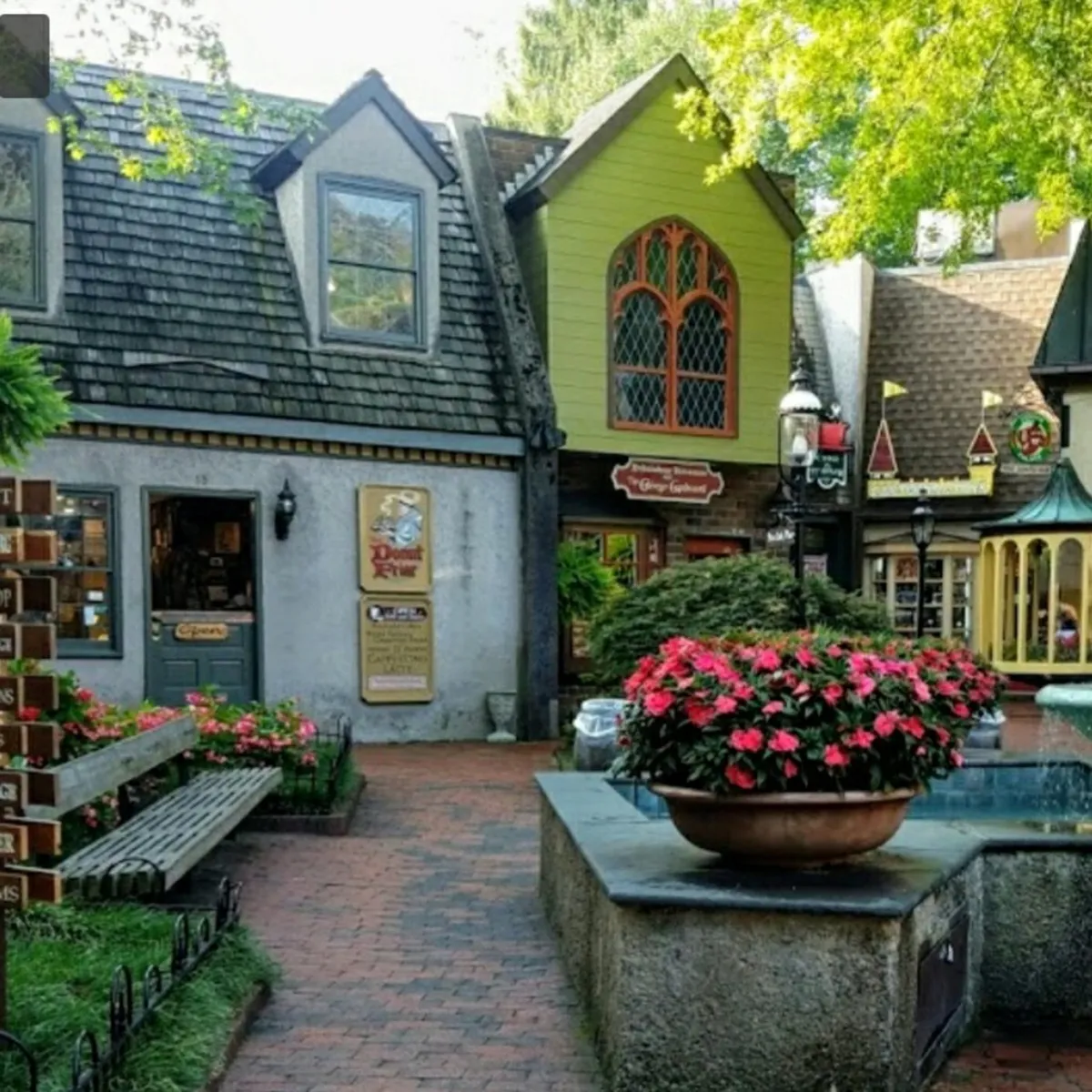 A lively cobblestone pathway lined with colorful storefronts, vibrant flower arrangements, and ornate lamp posts. The scene captures people strolling, shopping, and enjoying refreshments in a welcoming garden-like setting, evoking the charm of downtown Gatlinburg just steps from the condo.