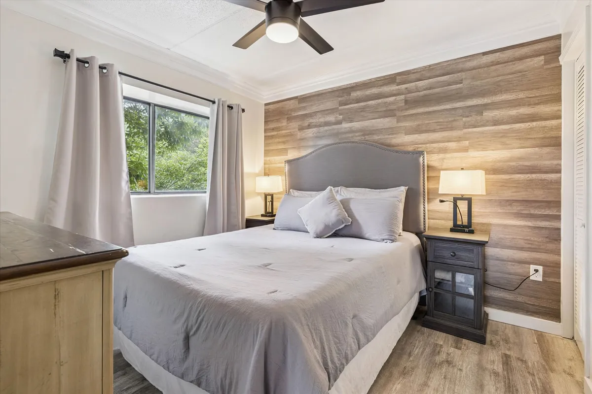 A cozy bedroom with a bed featuring a grey headboard and wooden accent wall. Table lamps provide soft lighting, and a window lets in gentle morning sunlight, creating a peaceful, welcoming space.