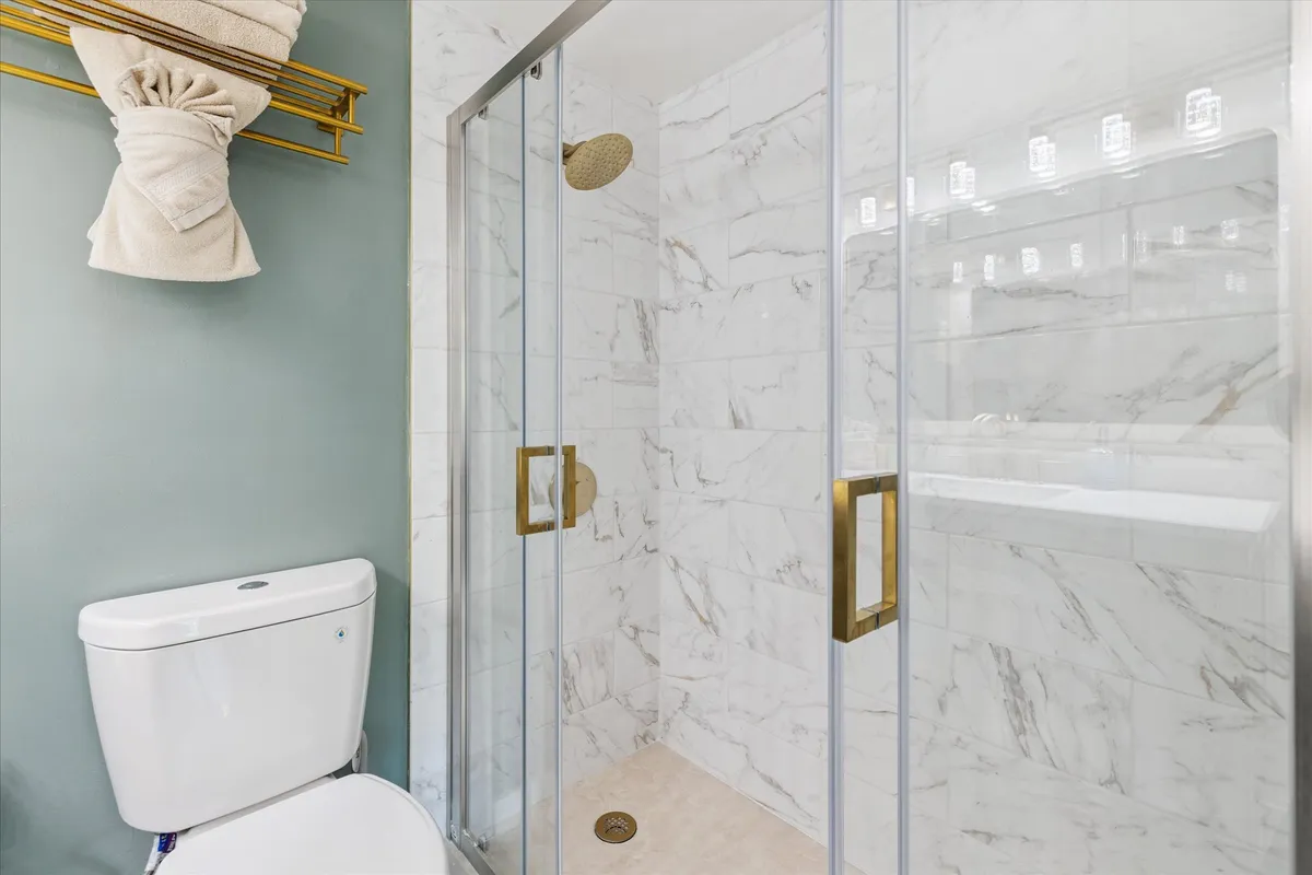 A stylish bathroom with a glass shower enclosure, gold fixtures, marble walls, and a contemporary toilet. The space exudes freshness and tranquility, ideal for relaxation and self-care.