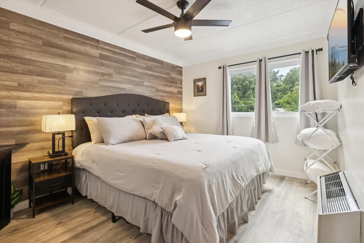 A king-sized bed with a tufted headboard is set against a wooden accent wall, flanked by a nightstand with a lamp. The room is bathed in natural light from a large window, creating a tranquil and refined atmosphere ideal for restful sleep.