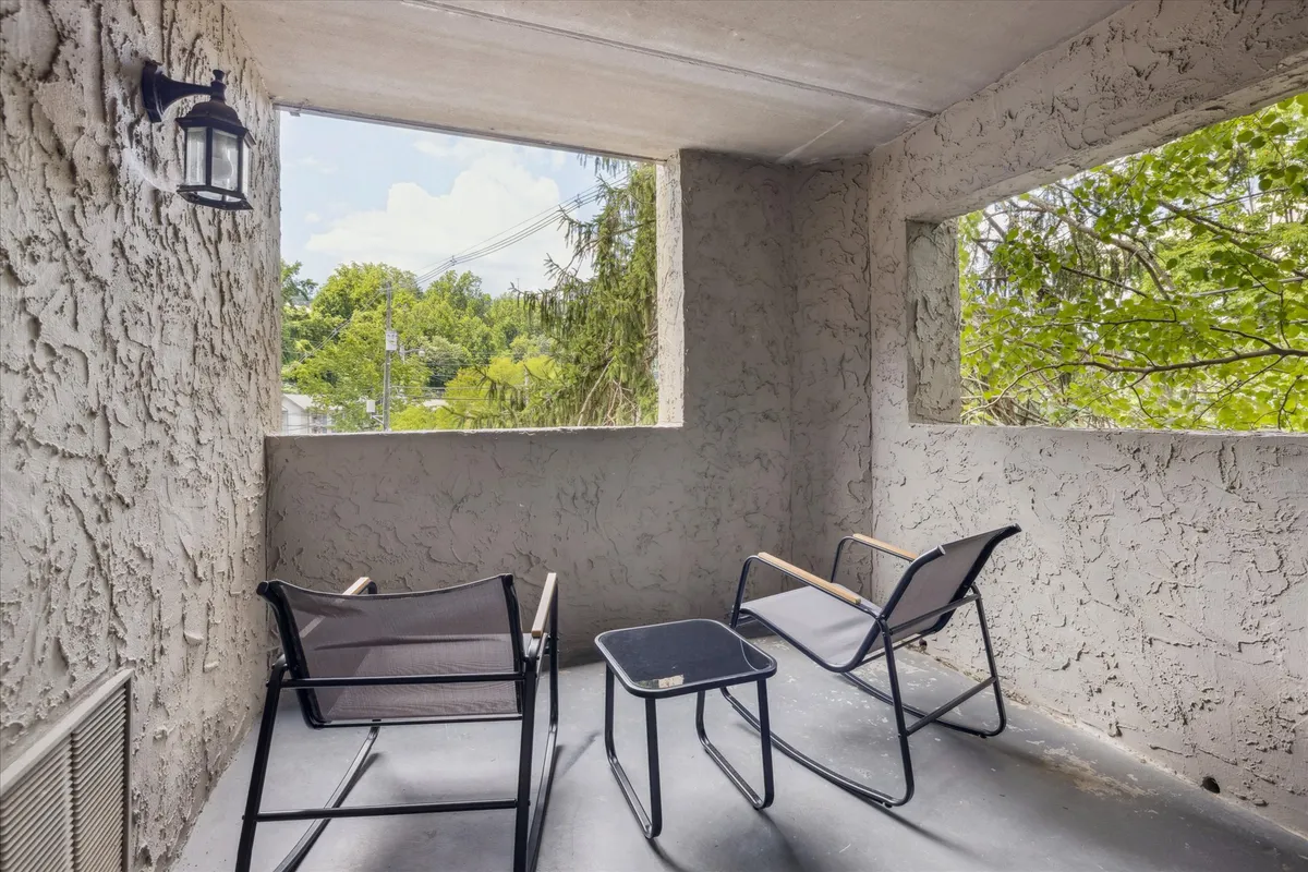 This image features two black chairs and a small table set by open windows with greenery visible outside. The arrangement suggests a tranquil spot for relaxing, reading, or enjoying a cool drink, evoking the leisure and comfort associated with poolside seating.