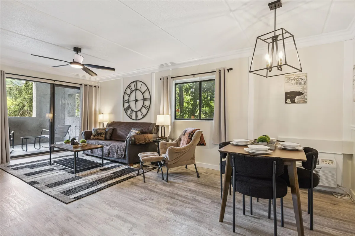 A sunlit open-concept space featuring a large circular wall clock, a dining table set for meals, a comfortable sofa and armchair, and expansive windows that fill the room with natural light. The layout allows for seamless movement between the dining and living areas, with contemporary touches throughout.