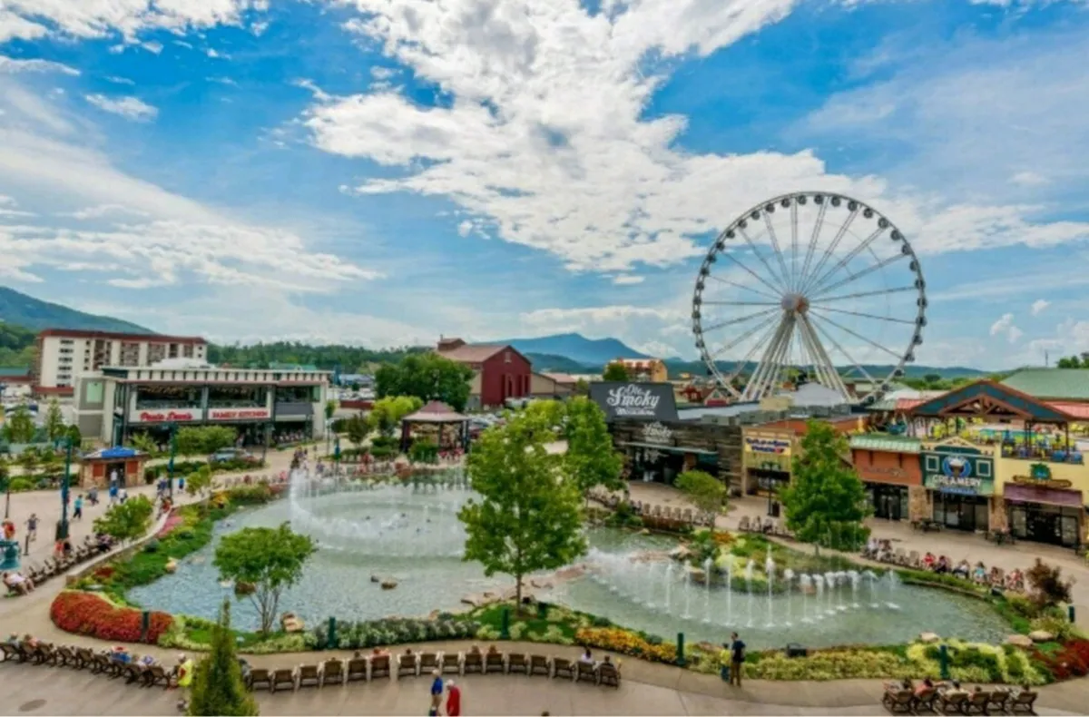 A lively outdoor scene highlighting a fountain, a ferris wheel, flower beds, and a bustling shopping and dining area—all packed with families enjoying the vibrant atmosphere. The image captures the spirit of fun and variety available on the main strip.