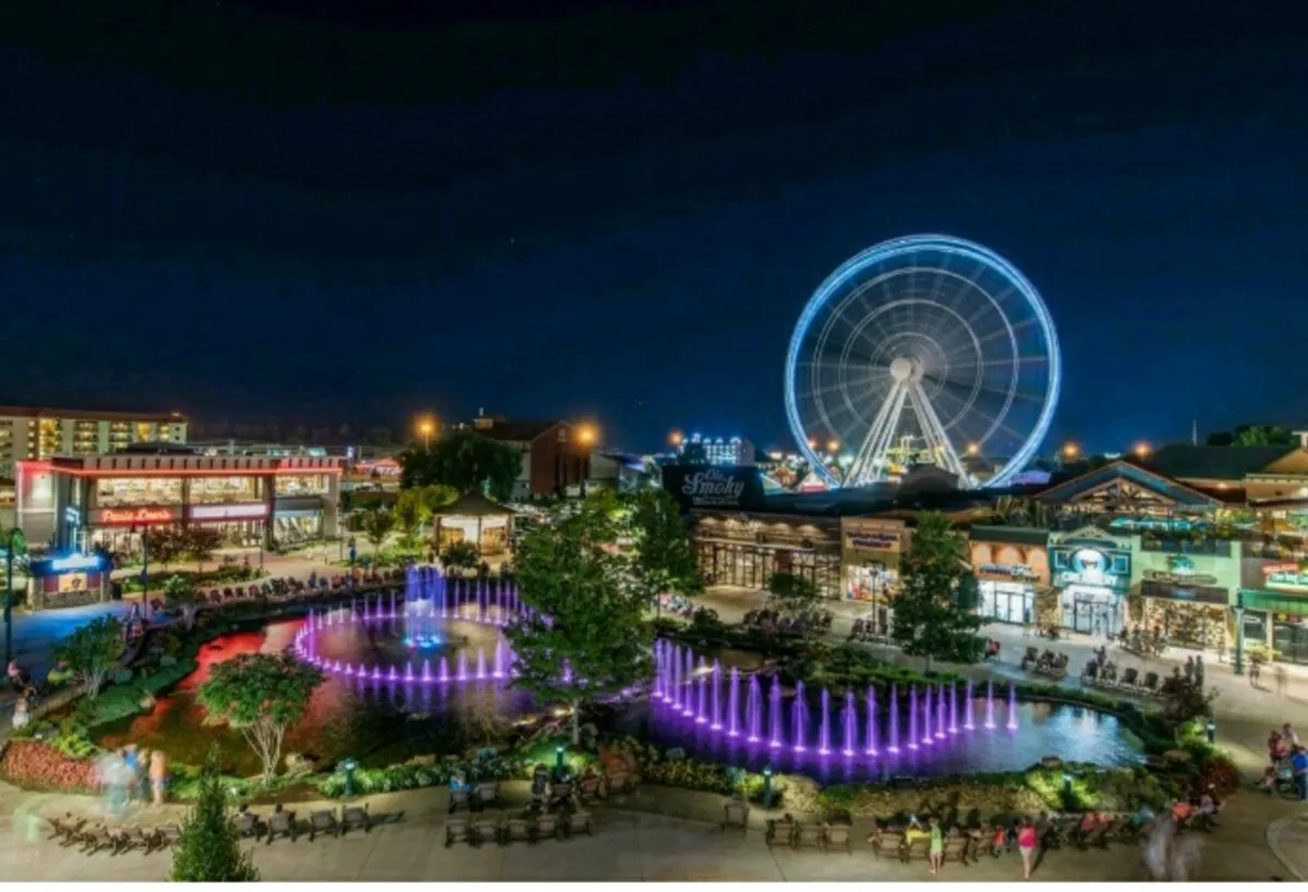 A lively outdoor plaza featuring a brightly lit Ferris wheel, colorful fountains, and a number of restaurants and shops. People are gathered around outdoor seating areas, enjoying the festive atmosphere and the impressive light show from the fountains.
