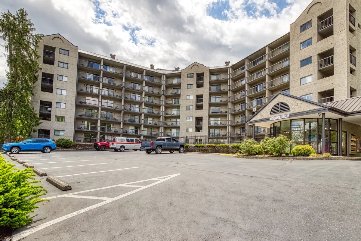 The hero image showcases a modern, multi-story condo building with a sleek glass entrance, surrounded by landscaped greenery. The parking lot in front offers easy access for guests, and balconies suggest opportunities for relaxation while overlooking sunset views. The overall mood is inviting and tranquil, hinting at community spaces and a central location near Gatlinburg's main attractions.